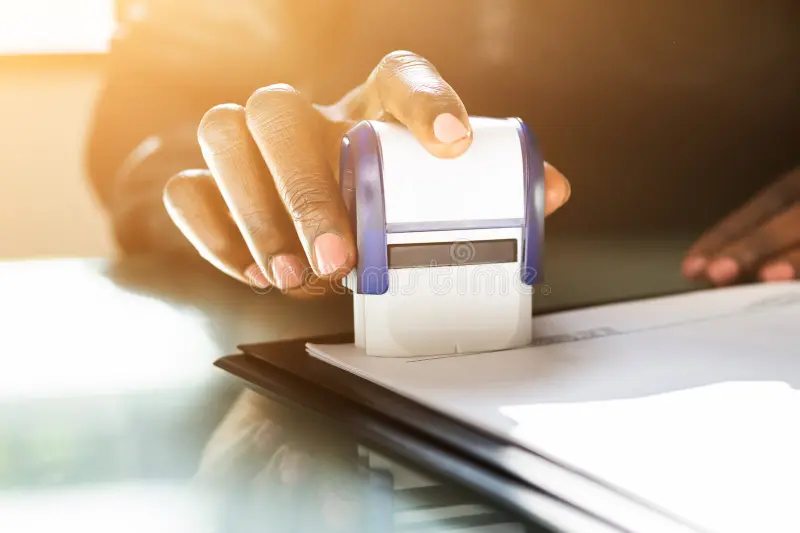 Hand pressing a rubber stamp onto a document on a desk in bright sunlight.
